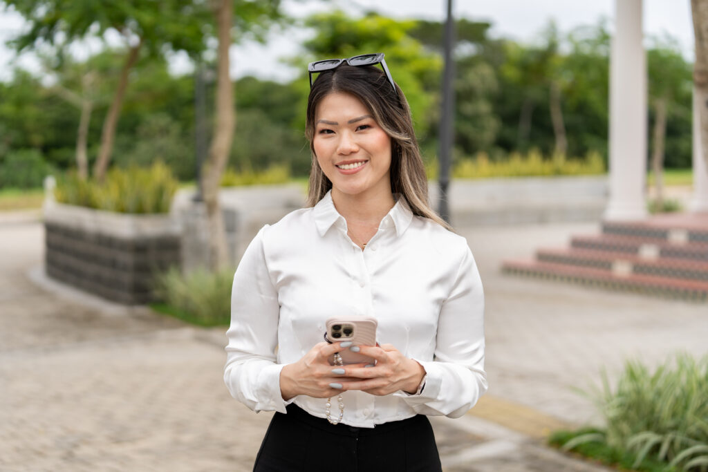 Mujer adulta sonrie a la camara mientras sostiene con sus dos manos su celular, de fondo esta la smart city de porta norte